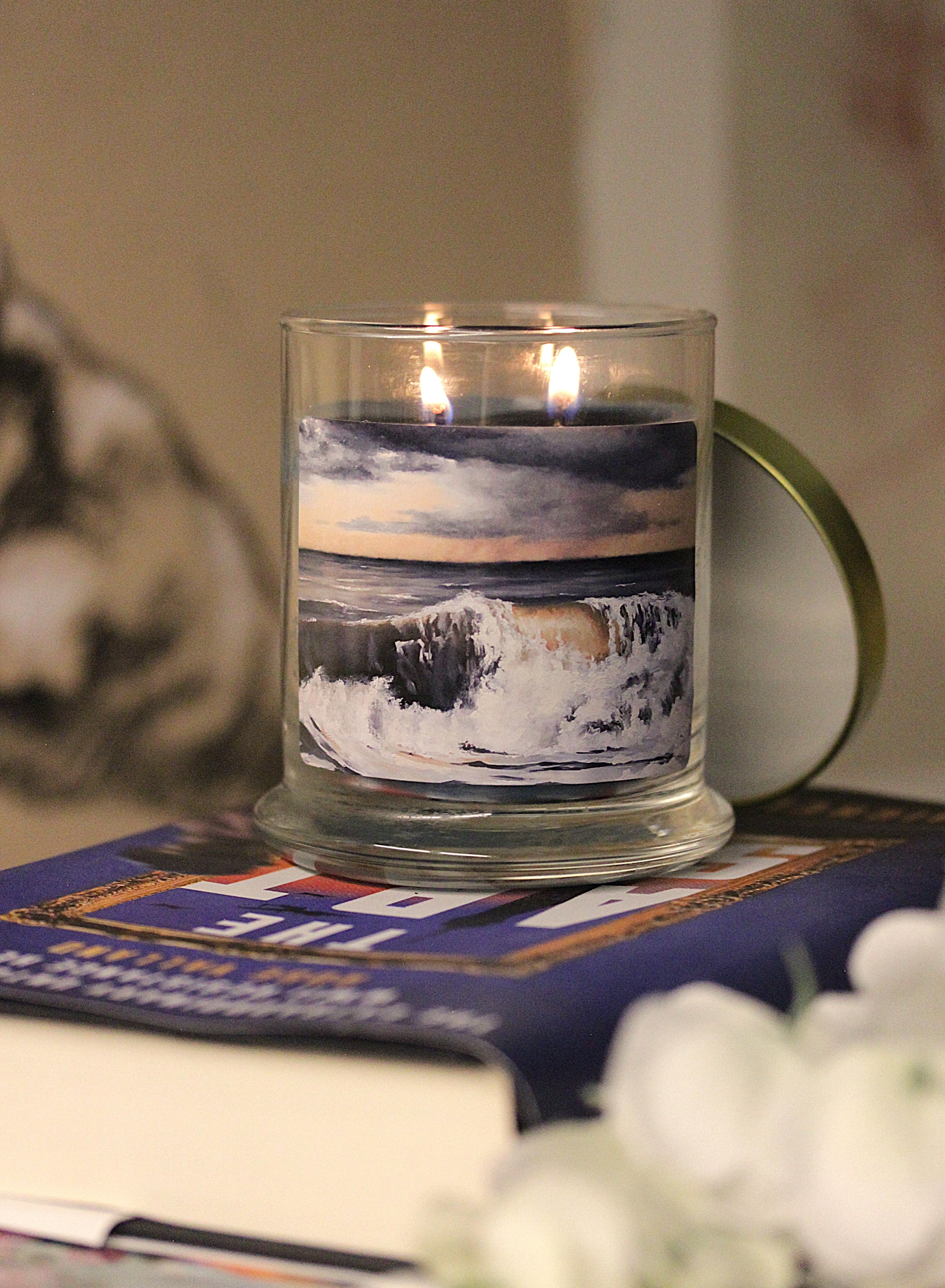 A lit 9oz sea salt scented candle with original coastal artwork on the glass jar in an aesthetic environment.