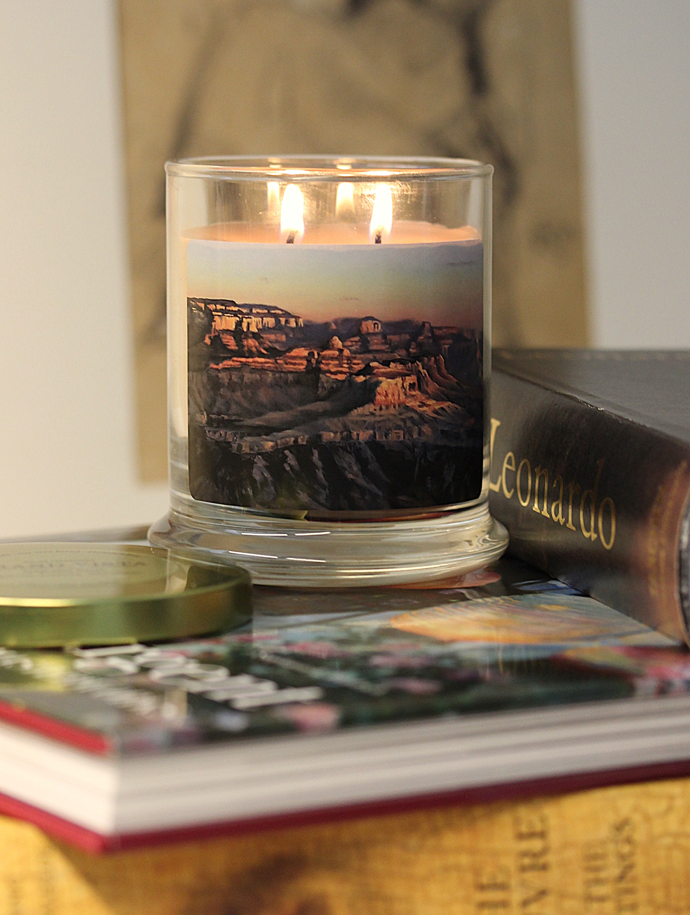A lit 9oz patchouli scented candle with original landscape artwork on the glass jar in an aesthetic environment.
