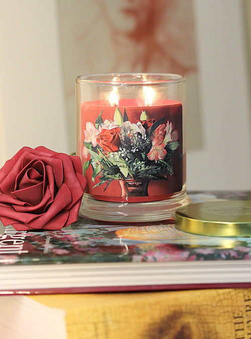a lit 9oz rose vanilla scented candle with original floral artwork on the glass jar in an aesthetic environment.